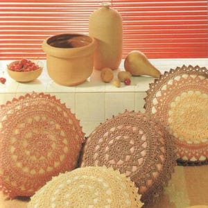 May include: Three crocheted round cushions in shades of beige, brown, and pink. The cushions have a delicate lace pattern and are arranged on a white tiled surface. There are small bowls of red cherries and other decorative items in the background.