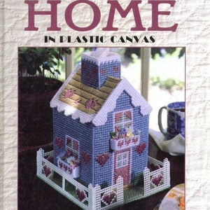 May include: A book cover with the title "No Place Like Home" in plastic canvas. The cover features a blue and white plastic canvas house with a white picket fence and a red door. The book cover also includes the text "Leisure Arts Presents" and "Over 75 Projects!"