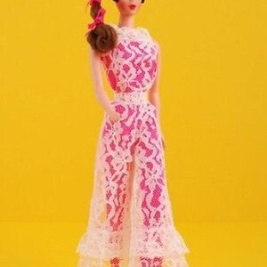 Può includere: Una bambola alla moda con capelli castani raccolti in una coda di cavallo laterale con nastri rosa, che indossa una tuta senza maniche con uno strato base rosa acceso coperto da un pizzo bianco con orlo a balze e scarpe col tacco rosa.