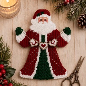 May include: A plastic canvas Santa Claus ornament featuring a red and green coat with white trim, a white beard, and a hat. The figure holds a string of three small, white, heart-shaped ornaments with red centers and gold-colored beaded edges.
