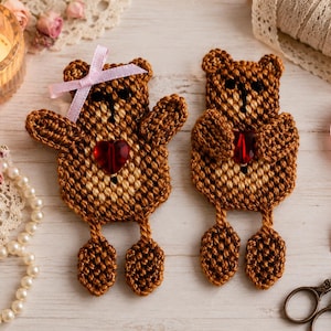 May include: Two brown crocheted teddy bear appliques with tan bellies and black stitched facial features. Each bear holds a small, faceted red heart-shaped bead. One bear features a light pink ribbon bow attached to its head.