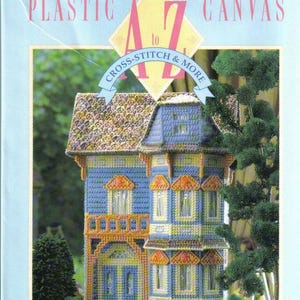 Peut inclure: Une couverture de livre avec le titre "Plastic Canvas A to Z Cross-Stitch & More" par Vanessa Ann's. La couverture présente un modèle de maison en toile plastique coloré avec un toit bleu et des bordures jaunes.