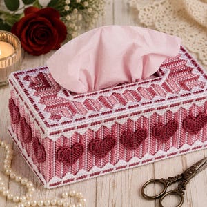 May include: A rectangular tissue box cover crafted from white and pink plastic canvas with a stitched geometric pattern and raised dark red heart motifs on the sides. A single light pink tissue is pulled through the top opening of the box.