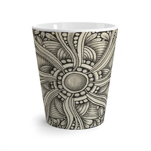 Könnte beinhalten: Eine weiße Keramiktasse mit einem schwarz-weißen abstrakten Design. Das Design zeigt einen zentralen Kreis mit einer grauen Mitte und einer Reihe konzentrischer Kreise und Linien, die nach außen strahlen.