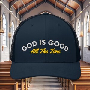 Könnte beinhalten: Marineblaue Trucker-Cap mit dem gestickten Text "GOD IS GOOD All The Time" in Weiß und Gelb. Die Kappe hat einen Mesh-Rücken und einen gebogenen Schirm. Der Hintergrund ist ein Kircheninnenraum.