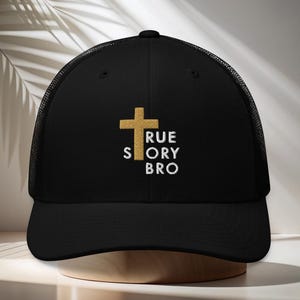 Könnte beinhalten: Schwarze Trucker-Cap mit einem goldenen Kreuz und dem gestickten Text "TRUE STORY BRO" in Weiß. Die Cap hat einen Mesh-Rücken und einen gebogenen Schirm. Die Cap wird auf einem Holzständer präsentiert.
