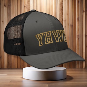 Op de afbeelding: Een grijze truckerpet met een zwarte mesh achterkant en een gebogen klep. De voorkant van de pet toont de gouden letters "YHW" in een lettertype in collegestijl. De pet wordt gepresenteerd op een witte sokkel.
