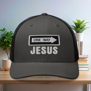 May include: A charcoal gray trucker hat with a black mesh back. The front of the hat features the text "ONE WAY" above an arrow, and the word "JESUS" below, all in white embroidered lettering.