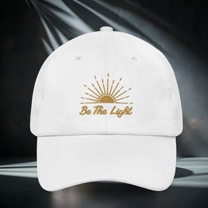 Könnte beinhalten: Weißes Baseballcap mit gebogenem Schirm. Die Kappe hat ein gesticktes Design einer Sonne mit Strahlen und dem Text "Be The Light" in Gold. Der Hut ist aus einem weichen Stoff gefertigt und hat einen verstellbaren Riemen.