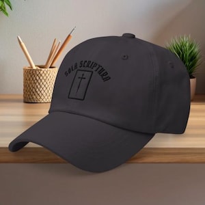 Puede incluir: Gorra de béisbol negra con la inscripción bordada "Sola Scriptura" sobre una cruz dentro de un rectángulo. La gorra tiene una visera curva y una corona estructurada. El sombrero se muestra sobre una superficie de madera.