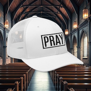 May include: White baseball cap with a mesh back and a curved brim. The word "PRAY" is printed in black, inside a black rectangle, on the front of the cap. The cap is set against a church interior background.