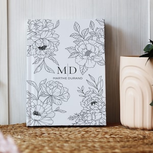 May include: A white journal with black floral line art. The cover features peony illustrations and the initials "MD" with the name "Marthe Durand" below. The journal is displayed on a woven surface.
