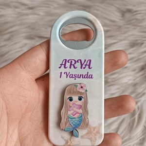 Puede incluir: Un abridor de botellas blanco con un dise&ntilde;o de sirena y el texto "ARYA 1 Yaşında".