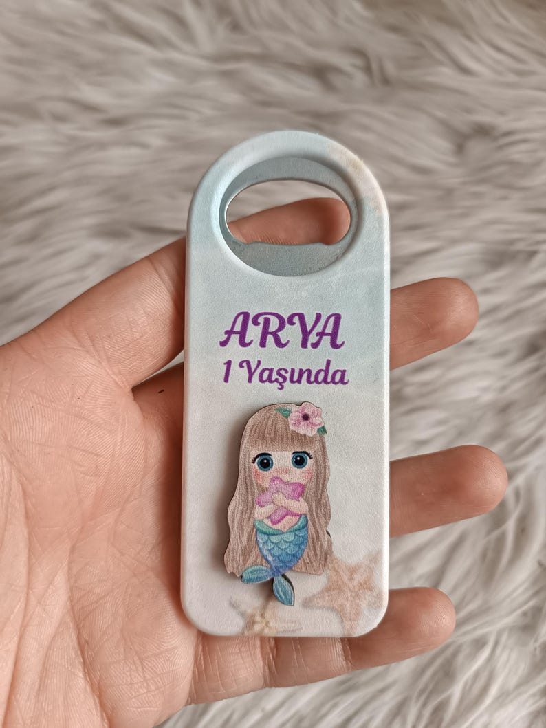 Puede incluir: Un abridor de botellas blanco con un dise&ntilde;o de sirena y el texto "ARYA 1 Yaşında".