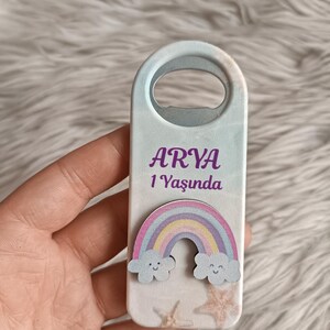 Puede incluir: Un abridor de botellas de madera blanco con un dise&ntilde;o de arco&iacute;ris y nubes. El abridor de botellas tiene el nombre "ARYA" y el texto "1 Yaşında" impreso en &eacute;l.