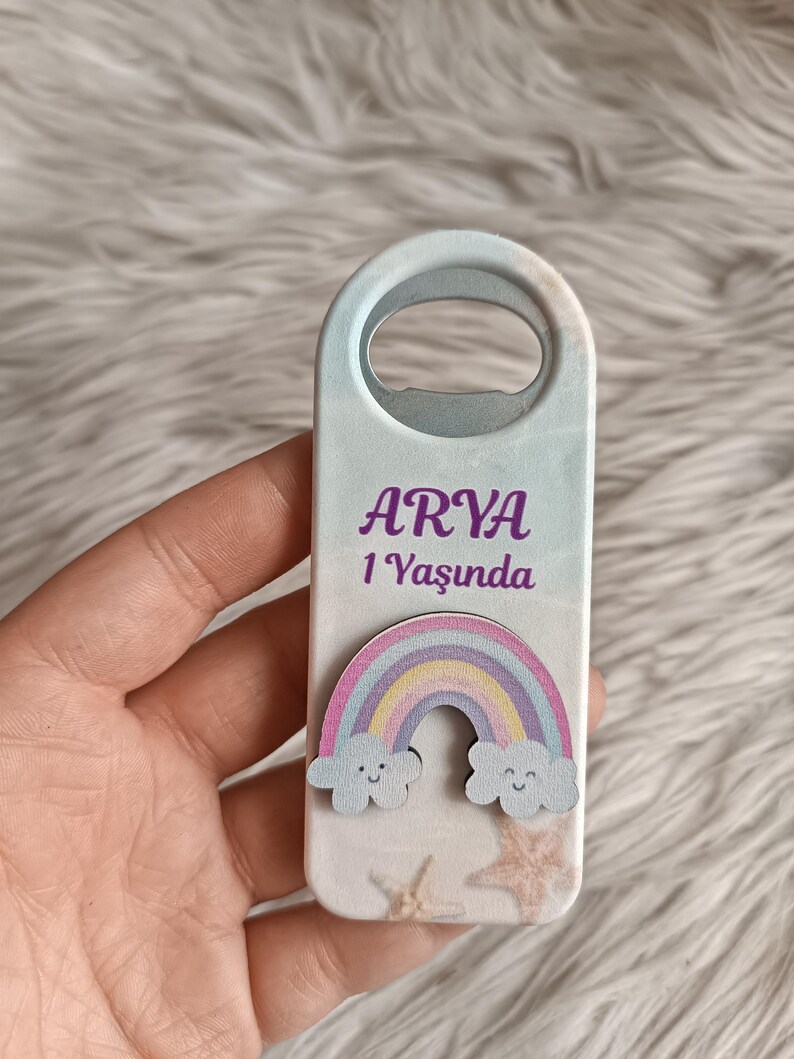Puede incluir: Un abridor de botellas de madera blanco con un dise&ntilde;o de arco&iacute;ris y nubes. El abridor de botellas tiene el nombre "ARYA" y el texto "1 Yaşında" impreso en &eacute;l.