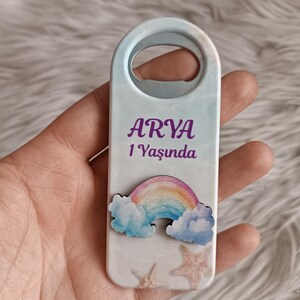 Puede incluir: Un abridor de botellas azul claro con un dise&ntilde;o de arco&iacute;ris y nubes. El abridor de botellas tiene el nombre "ARYA" y el texto "1 Yaşında".