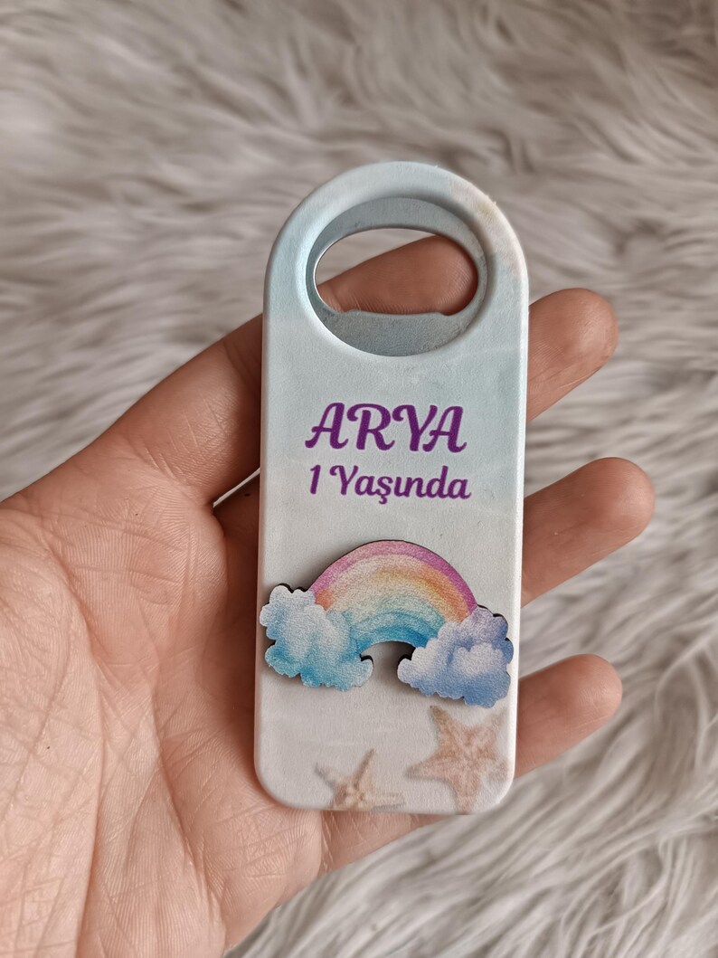 Puede incluir: Un abridor de botellas azul claro con un dise&ntilde;o de arco&iacute;ris y nubes. El abridor de botellas tiene el nombre "ARYA" y el texto "1 Yaşında".