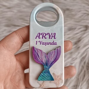 Puede incluir: Un abridor de botellas blanco con un dise&ntilde;o de cola de sirena. El abridor de botellas tiene una cola de sirena morada y turquesa sobre un fondo azul claro. El texto "ARYA 1 Yaşında" est&aacute; impreso en el abridor de botellas.