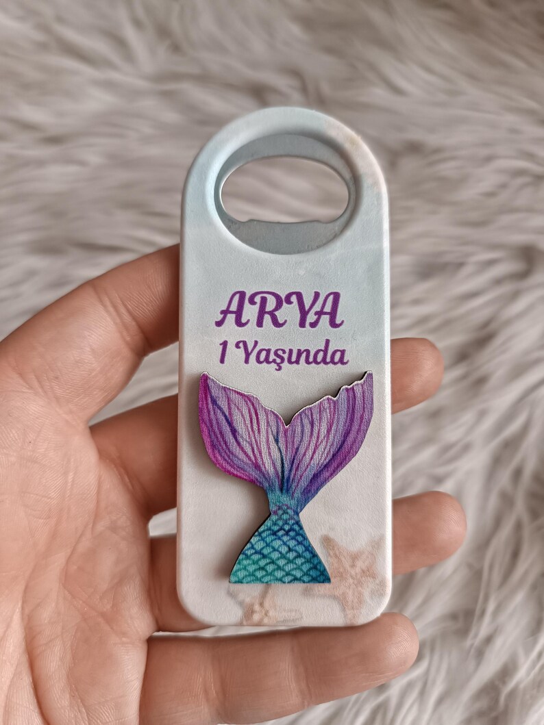 Puede incluir: Un abridor de botellas blanco con un dise&ntilde;o de cola de sirena. El abridor de botellas tiene una cola de sirena morada y turquesa sobre un fondo azul claro. El texto "ARYA 1 Yaşında" est&aacute; impreso en el abridor de botellas.