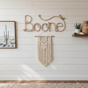 May include: A wall-mounted decor arrangement featuring a rope sign spelling "Boone" above a cream-colored macrame wall hanging. A framed black and white desert landscape and a small shelf with plants and a basket complete the scene.