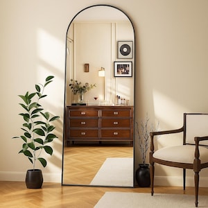 May include: A full-length arched mirror with a black frame. The mirror reflects a room with a wooden dresser, a plant in a black pot, and a chair. Two framed pictures hang on the wall. The room has a neutral color palette.
