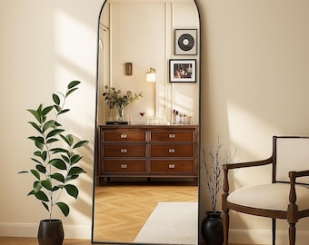 May include: A full-length arched mirror with a black frame. The mirror reflects a room with a wooden dresser, a plant in a black pot, and a chair. Two framed pictures hang on the wall. The room has a neutral color palette.