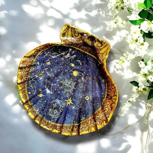Könnte beinhalten: Dekorative Muschelschalen-Schmuckschale. Die Schale hat ein tiefblaues Inneres mit einem Himmelsdesign aus goldenen Sternen, Monden und Sternbildern. Der Rand ist mit einem goldenen und braunen Zierornament verziert, was ein einzigartiges Kunstwerk schafft.