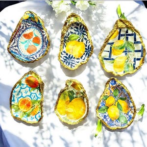 May include: Six decorative oyster shell dishes with gold trim and various lemon and citrus fruit designs. The dishes are on a white surface.