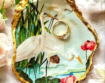 Può includere: Un portagioie decorativo con bordo dorato e interno dipinto raffigurante un airone bianco, fiori rossi e fogliame verde. Un anello d'oro con una grande pietra trasparente è all'interno. Il piatto ha una forma ovale irregolare.