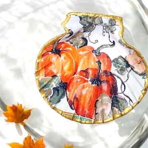 Puede incluir: Un plato decorativo en forma de concha de vieira con un diseño pintado de calabazas de color naranja brillante con enredaderas y hojas verdes. Los bordes de la concha están rematados con pintura dorada metálica, creando un borde decorativo.