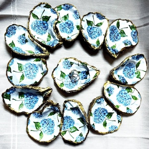 Puede incluir: Doce pequeños platos en forma de concha de ostra blancos con borde dorado. Cada plato está decorado con un patrón de flores de hortensia azules. Un plato tiene dos anillos plateados en su interior.