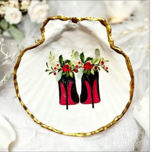 May include: A white seashell-shaped decorative dish with a gold-painted rim. The dish features a painted design of black and red high-heeled shoes adorned with floral arrangements of red berries and green leaves.
