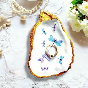 Pode incluir: Um prato decorativo feito de concha de ostra com interior branco, decorado com ilustrações de libélulas azuis e roxas e uma borda pintada de dourado. Um anel dourado com uma pedra transparente repousa no centro da concha.
