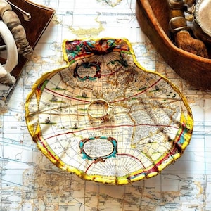 Peut inclure: Un plat décoratif en forme de coquillage avec un motif de carte vintage. Le plat a un bord doré et une impression de carte détaillée dans des tons beige, rouge et bleu. Une bague dorée repose au centre du plat, posé sur une carte.