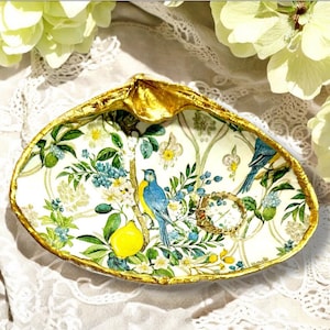 May include: A decorative seashell trinket dish with a gold-painted rim. The interior features a botanical design with blue birds, lemons, and floral elements. A gold-toned ring rests inside the dish, which is set on a white lace surface.