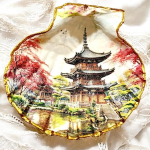 以下が含まれることがあります： 日本の塔と周囲の風景が描かれた装飾的な貝殻。 赤と緑の木々、池、金色の縁取りが特徴です。 貝殻は白く、質感のある表面に置かれています。
