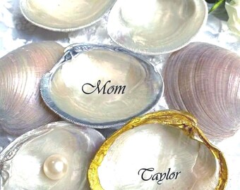 Puede incluir: Una colección de conchas de almeja blancas pulidas, algunas con bordes pintados en oro o plata metálicos. Una concha tiene inscrita la palabra Mom, otra el nombre Taylor, y una concha contiene una perla blanca única en su centro.