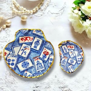 Può includere: Due piattini decorativi a forma di conchiglia con un motivo blu e bianco di tessere del mahjong con accenti rossi e blu. Ogni piattino presenta un bordo smerlato dipinto in oro, con illustrazioni di draghi, vasi e motivi floreali.