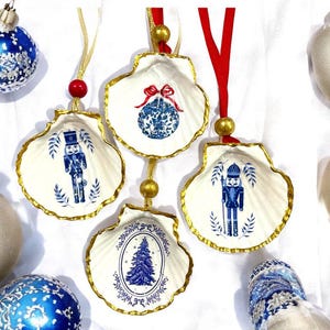 Peut inclure: Ornements de Noël en coquillage blanc avec bordure dorée et motifs bleus et blancs. Les ornements présentent des images d'un casse-noisette, d'un sapin de Noël et d'une boule décorée. Chaque ornement est suspendu à un ruban rouge ou doré.
