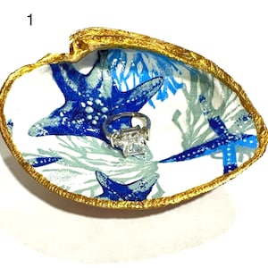 May include: A white seashell ring dish with a gold rim. The dish is decorated with a blue and white starfish and coral design. A ring is sitting in the center of the dish.