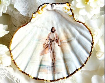 Puede incluir: Una concha marina blanca decorativa con un borde pintado de oro. El interior presenta una imagen pintada de una figura religiosa con túnicas fluidas. La concha está rodeada de flores blancas y encaje.
