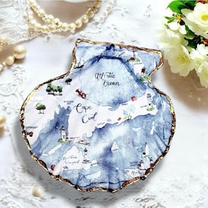 Puede incluir: Una concha decorativa con un mapa en acuarela azul y blanco de Cape Cod y el Océano Atlántico. La concha tiene un borde festoneado pintado en dorado e incluye ilustraciones de faros, árboles y una ballena con etiquetas de texto impresas.