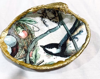 Può includere: Una conchiglia di ostrica decorativa con un'illustrazione a decoupage di una cincia dal ciuffo appollaiata su un ramo vicino a un nido con uova blu. La superficie interna è bianca con un motivo di uccelli e il bordo è dipinto in oro.