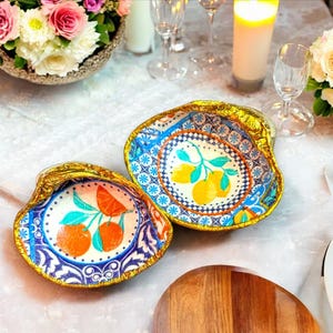 Pode incluir: Duas tigelas decorativas de cerâmica em forma de concha com uma borda dourada. As tigelas são decoradas com um padrão azul e branco e apresentam um motivo de frutas cítricas com limões e laranjas.