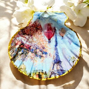 Puede incluir: Un plato decorativo de concha con una pintura de la Torre Eiffel y globos aerostáticos. La concha tiene un borde dorado y un diseño artístico colorido, sobre un cielo azul.