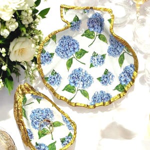 May include: Two shell-shaped dishes with a white base, featuring a blue hydrangea floral pattern and textured gold-painted edges. The larger dish is scallop-shaped, while the smaller dish is oyster-shaped and holds a thin gold ring.