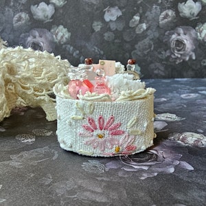 May include: A small white fabric basket with pink and white embroidered flowers. The basket is filled with small glass bottles with cork tops and white cotton balls.