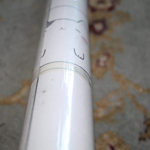 May include: A rolled-up white paper product with a repeating pattern of a cartoon cat face. The cat face has black whiskers, eyes, and a pink nose. The paper is wrapped in clear plastic.