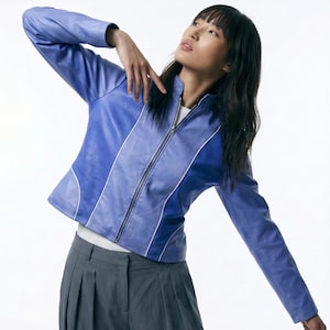 以下が含まれることがあります： 白のストライプとシルバーのジッパーが付いた、ペリウィンクルブルーのジャケット。 ジャケットは長袖で、スタンドカラーです。 モデルはグレーのプリーツパンツを着用しています。